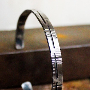 May include: A silver bracelet with a geometric design. The bracelet is made of a thin metal band with a series of parallel lines etched into the surface.