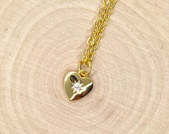 May include: A gold heart-shaped pendant necklace with a small sparkling stone in the center. The necklace is on a wooden surface.