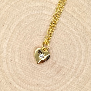 Può includere: Una collana con un ciondolo a forma di cuore in oro con un piccolo sasso scintillante al centro. La collana è su una superficie di legno.
