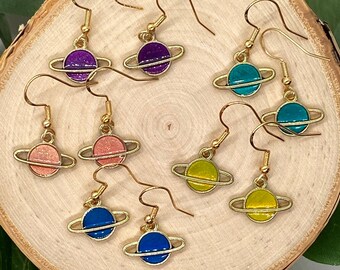 May include: A set of eight gold-toned earrings featuring a small planet design with a ring around it. The planets are in various colors, including purple, teal, pink, yellow, and blue.