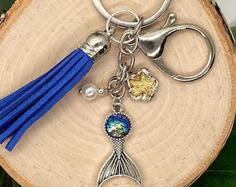 May include: A blue tassel keychain with a silver mermaid tail charm, a pearl bead, and a gold starfish charm. The keychain is attached to a silver key ring.