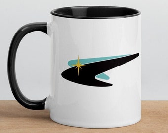 May include: White ceramic mug with a black handle and interior. The mug features a retro-style design with a black, curved shape, a light blue accent, and a gold starburst. The mug is sitting on a wooden surface.