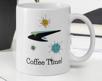 May include: White ceramic mug with a mid-century modern design featuring a black, teal, and yellow graphic with the words "Coffee Time!" printed in black. The mug is on a white surface with a keyboard and a plant in the background.