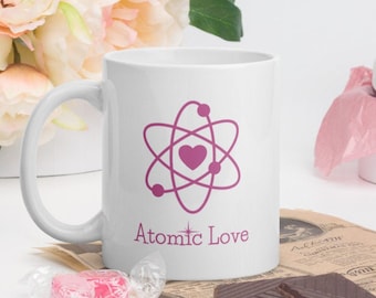 May include: White ceramic mug with a pink atom symbol and a pink heart in the center. The text "Atomic Love" is printed below the symbol.