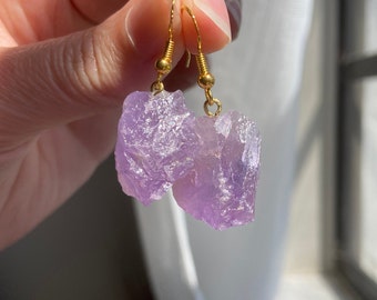 May include: A pair of amethyst crystal earrings with gold hooks. The amethyst crystals are raw and have a natural, irregular shape.