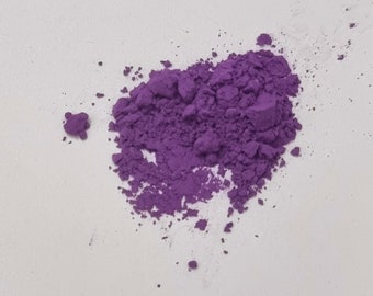 Puede incluir: Un pequeño montón irregular de polvo de pigmento morado vibrante. La sustancia tiene una textura seca y granular con grumos de varios tamaños dispersos por la superficie, creando un montículo concentrado de color violeta intenso.