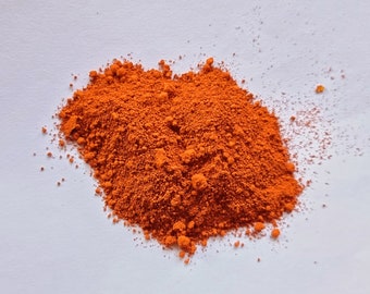 May include: A pile of bright orange powder on a white surface. The powder is finely ground and has a smooth texture.