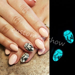 May include: Close-up of fingernails painted a light pink colour with black and white dreamcatcher designs. The nails are glowing a bright blue in the dark.
