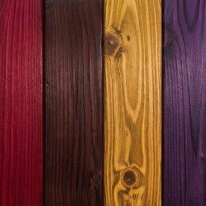 Puede incluir: Seis tablones de madera verticales dispuestos uno al lado del otro, cada uno con un acabado teñido distinto. Los colores incluyen marrón claro natural, rojo intenso, marrón oscuro, amarillo dorado, morado oscuro y rosa vibrante, mostrando la veta.