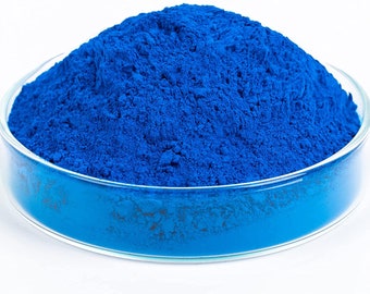 May include: A close-up of a pile of bright blue powder in a clear glass dish. The powder is finely ground and has a smooth, uniform texture.