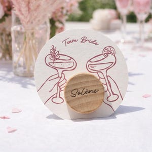 Può includere: Un segnaposto rotondo per matrimoni bianco sporco con un dettaglio in legno. La carta presenta un disegno a tratto di due mani che tengono bicchieri da cocktail, il testo "Team Bride" e il nome "Solène". Fiori e bicchieri rosa sullo sfondo.
