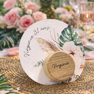 Può includere: Un segnaposto rotondo in stile acquerello con disegni floreali e di piume. La carta ha il nome "Virginie" e il testo "31 ans". Un piccolo pezzo di legno con il nome "Virginie" è attaccato.