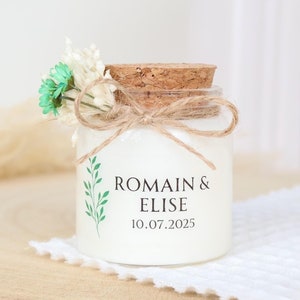 Puede incluir: Un pequeño frasco de vidrio con tapa de corcho y una etiqueta blanca. La etiqueta tiene hojas verdes y el texto "ROMAIN & ELISE 10.07.2025". El frasco está decorado con un pequeño ramo de flores blancas y verdes y un lazo de cuerda marrón.