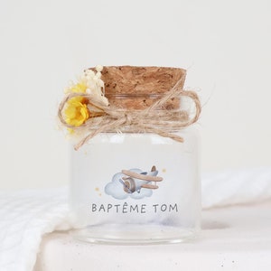 Può includere: Un piccolo barattolo di vetro con un tappo di sughero, decorato con un aereo blu e nuvole. Il barattolo ha un'etichetta che dice "BAPTÊME TOM". Il barattolo è legato con uno spago e decorato con fiori gialli secchi.