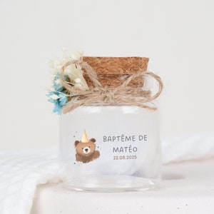 Può includere: Un piccolo barattolo di vetro con un tappo di sughero, decorato con fiori secchi e un'etichetta che recita "Baptême de Mateo 22.08.2025". L'etichetta presenta anche un orso dei cartoni animati con un cappello da festa.