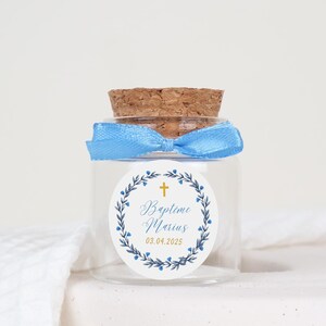 Peut inclure: Un petit pot en verre avec un bouchon en liège et un ruban bleu noué autour du cou. Le pot a un autocollant blanc avec une couronne de laurier bleue, une croix dorée et le texte "Baptime Marius 03.04.2025".