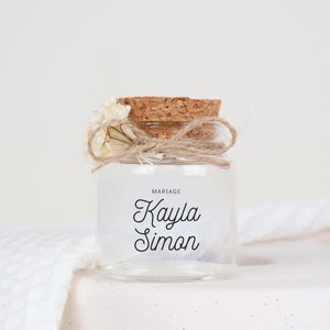 Può includere: Un piccolo barattolo di vetro con un tappo di sughero e un fiocco di spago. Il barattolo è pieno di fiori secchi e ha la scritta "MARIAGE Kayla Simon" stampata sul davanti.