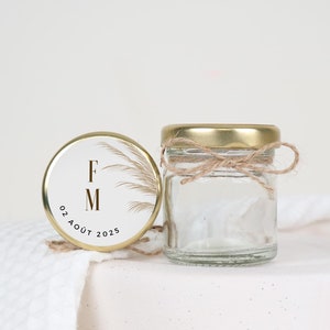 Può includere: Un piccolo barattolo di vetro trasparente con coperchio dorato e spago legato attorno al collo. Accanto, un coperchio abbinato con le iniziali "FM", la data "02 AOÛT 2025" e un design botanico. Il barattolo è su una superficie bianca.