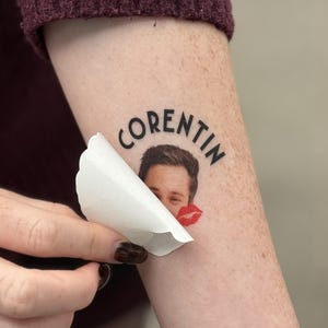 Peut inclure: Un tatouage temporaire avec le nom "CORENTIN" en texte noir arqué. Le tatouage présente une photo d'un visage et une marque de baiser rouge. Le papier de support est en train d'être décollé.