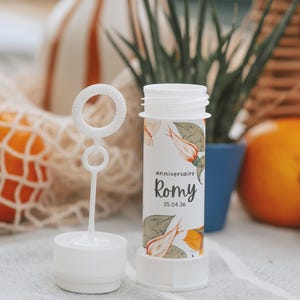 Könnte beinhalten: Ein weißer Seifenblasenstab aus Kunststoff mit einem botanischen Aufdruck aus orangefarbenen und grünen Blättern. Das Etikett zeigt den Text "anniversaire Romy 25.04.26" in einer dunklen Schreibschrift. Der Behälter ist zylindrisch mit Schraubverschluss.