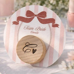 Può includere: Un biglietto rotondo a righe rosa con la scritta "Team Bride Camille" e un disco di legno con un cuore e un'iniziale. Il biglietto presenta un motivo a fiocco e nastro rosa. Una candela accesa, smalto per unghie e fiori sono sullo sfondo.