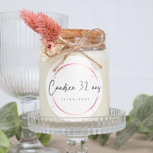 Może przedstawiać: Biała świeca w szklanym słoiku z korkową pokrywką. Słoik ma okrągłą etykietę z różowym i złotym wzorem. Etykieta zawiera napis "Candice 32 ans 11/02/2025".