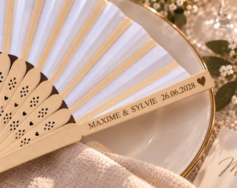 Puede incluir: Un abanico plegable con tela blanca y varillas de madera clara. La estructura de madera presenta patrones de corazones y flores cortados con láser. La varilla exterior está grabada con el texto MAXIME & SYLVIE, la fecha 26.06.2028 y un corazón.