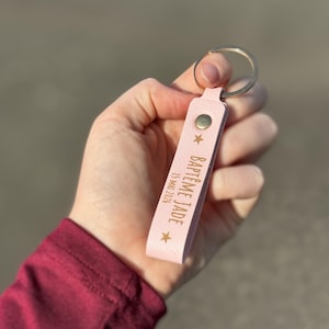 Peut inclure: Un porte-clés en cuir rose avec un anneau argenté. Le porte-clés porte l'inscription "BAPTÊME JADE 15 MAI 2026" en lettres dorées, ainsi que deux étoiles dorées. Un bouton-pression doré sécurise la lanière.