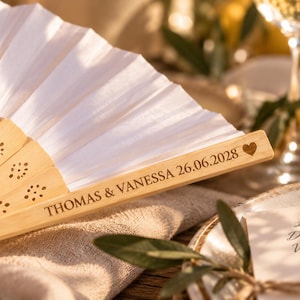 Puede incluir: Un abanico plegable con tela blanca y varillas de madera clara. La varilla exterior de madera está grabada con el texto THOMAS & VANESSA 26.06.2028 y un pequeño símbolo de corazón. Las varillas de madera presentan un patrón floral recortado.