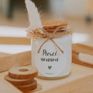 Può includere: Una candela bianca in un barattolo di vetro con coperchio in sughero, legata con spago e una piuma decorativa. L'etichetta recita "Merci nounou" con un cuore verde e il nome "Gabriel". La candela è su un vassoio di legno con sottobicchieri di legno.