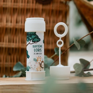 Peut inclure: Bouteille de bulles blanche avec un motif sur le thème de la jungle, avec un éléphant, une girafe et un lion. La bouteille porte l'inscription "BAPTÊME LORIS 26 JUIN 2026". Une baguette à bulles blanche est à côté de la bouteille.