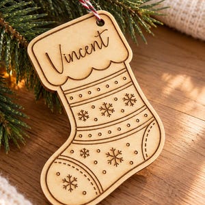 Può includere: Un addobbo natalizio in legno chiaro a forma di calza, con il nome Vincent inciso in corsivo sulla parte superiore. La calza è decorata con motivi a fiocco di neve e piccoli punti tagliati al laser, ed è appesa a uno spago rosso e bianco.
