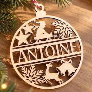 Op de afbeelding: Een cirkelvormig ornament van lichtgekleurd hout met een lasergesneden ontwerp en de naam ANTOINE in het midden. Het bovenste gedeelte toont een persoon op een slee en een sneeuwvlok, terwijl het onderste gedeelte een rendier en twee klokken bevat.