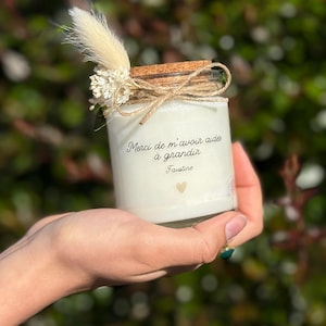 Op de afbeelding: Een witte kaars in een glazen pot met een kurken deksel, vastgebonden met touw en versierd met gedroogde bloemen. De kaars heeft de tekst "Merci de m'avoir aidée à grandir Faustine" en een klein gouden hart.