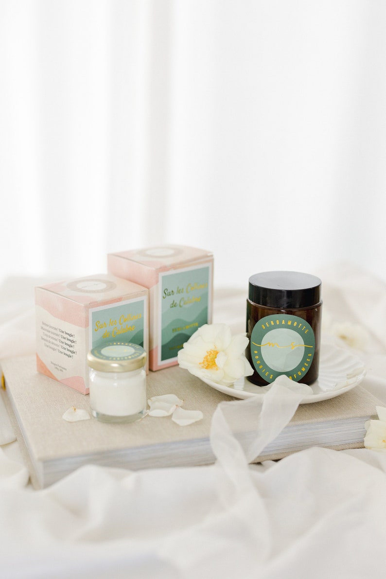 May include: Two pink and green boxes with the text "Sur les Collines de Calabre" and a small glass jar with a white candle. A white rose sits on a white plate next to a brown glass jar with a green label that reads "Bergamotte Bougies & Parfums".