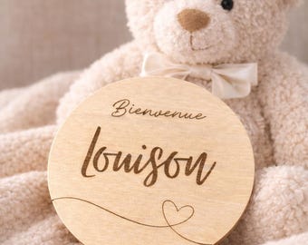 May include: A plush, cream-colored teddy bear with a cream bow tie, holding a round wooden sign. The sign has the text "Bienvenue Louison" in a script font, with a heart design. The bear is on a cream-colored blanket.