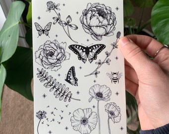 Può includere: Un'illustrazione in bianco e nero di fiori, farfalle e un'ape. L'illustrazione è su uno sfondo bianco con una texture sottile.
