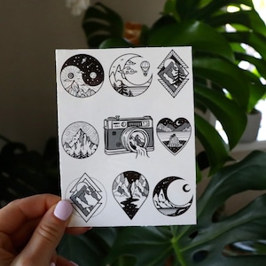 Peut inclure: Une feuille de tatouages temporaires noirs et blancs avec neuf motifs différents. Les motifs comprennent un croissant de lune, des montagnes, un ballon à air chaud, un appareil photo et un cœur.