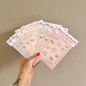 May include: A hand holds a fan of invitation cards with a beach theme. Each card has a scalloped edge and features the text "Gracie's Bachelorette, Traverse City, MI." and beach-related illustrations in red. The date "June 2026" is also visible.