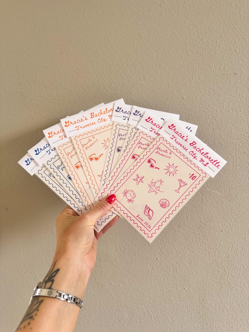 May include: A hand holds a fan of invitation cards with a beach theme. Each card has a scalloped edge and features the text "Gracie's Bachelorette, Traverse City, MI." and beach-related illustrations in red. The date "June 2026" is also visible.