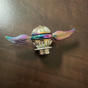 May include: A gold and iridescent metal spinning top with wings. The wings are iridescent and have a purple and gold color. The top is a sphere with a ring around the middle. The top is on a brown surface.