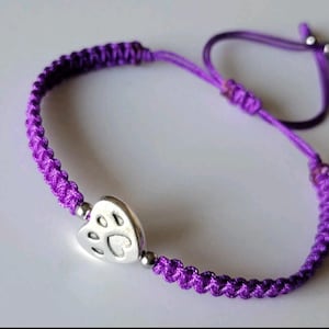 May include: A purple braided cord bracelet featuring a silver-toned heart-shaped charm with an embossed paw print design. The bracelet includes small silver-toned beads on either side of the charm and an adjustable sliding knot closure mechanism.