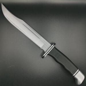 Könnte beinhalten: Ein silbernes Jagdmesser mit schwarzem Griff und silberner Klinge. Die Klinge hat eine gezahnte Kante und eine spitze Spitze.