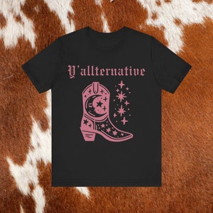 Puede incluir: Camiseta negra con el texto "Y'allternative" en letras rosas. Debajo del texto hay una bota vaquera rosa con una luna creciente y estrellas.