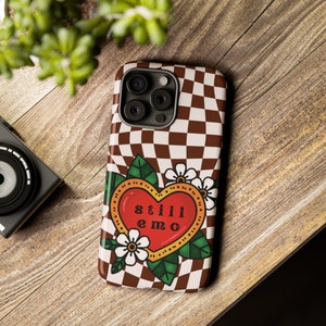 May include: A brown and white checkered phone case with a red heart and the text "still emo" in a yellow outline. The heart is surrounded by green leaves and white flowers.