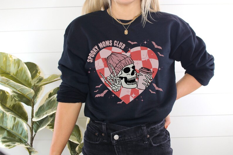 Peut inclure: Sweat-shirt noir avec un graphique de c&oelig;ur rose et rouge repr&eacute;sentant un squelette portant un bonnet rose et tenant une tasse de caf&eacute;. Le texte "Spooky Moms Club" est &eacute;crit en blanc sur le c&oelig;ur.