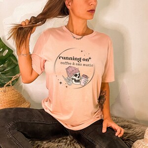 May include: A pink t-shirt featuring a graphic of a skeleton wearing a pink beanie and holding a coffee cup. The graphic is enclosed in a circle with the text "running on coffee & emo music".