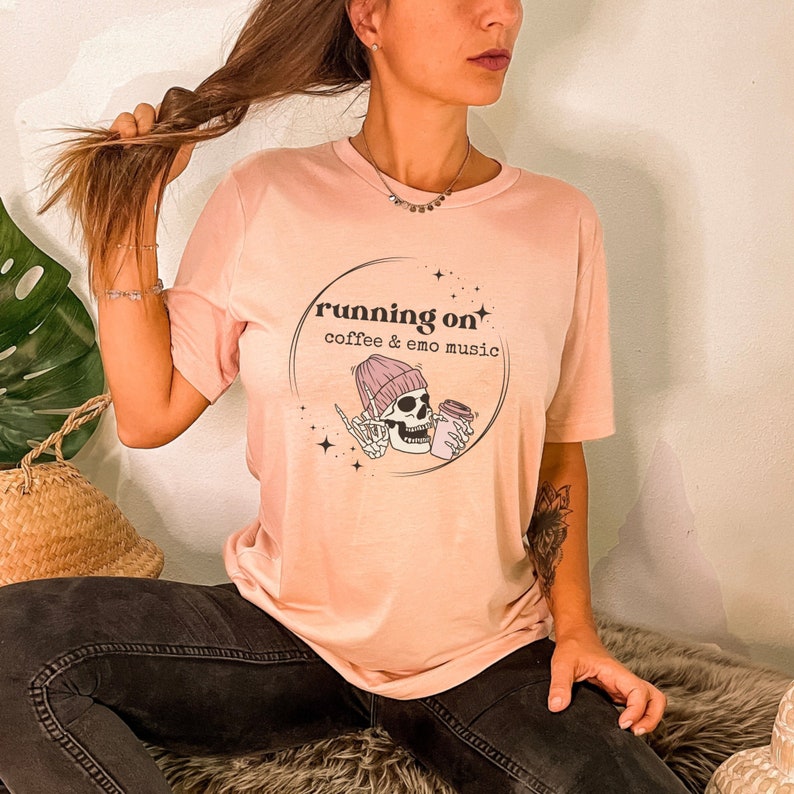 May include: A pink t-shirt featuring a graphic of a skeleton wearing a pink beanie and holding a coffee cup. The graphic is enclosed in a circle with the text "running on coffee & emo music".