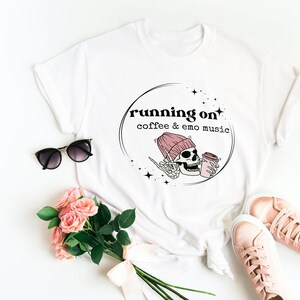 May include: White t-shirt with a black and pink graphic design. The graphic features a skull wearing a pink beanie and holding a cup of coffee. The text "running on coffee & emo music" is written in black above the skull.