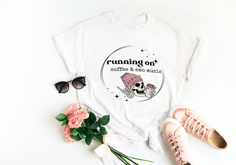 May include: White t-shirt with a black and pink graphic design. The graphic features a skull wearing a pink beanie and holding a cup of coffee. The text "running on coffee & emo music" is written in black above the skull.